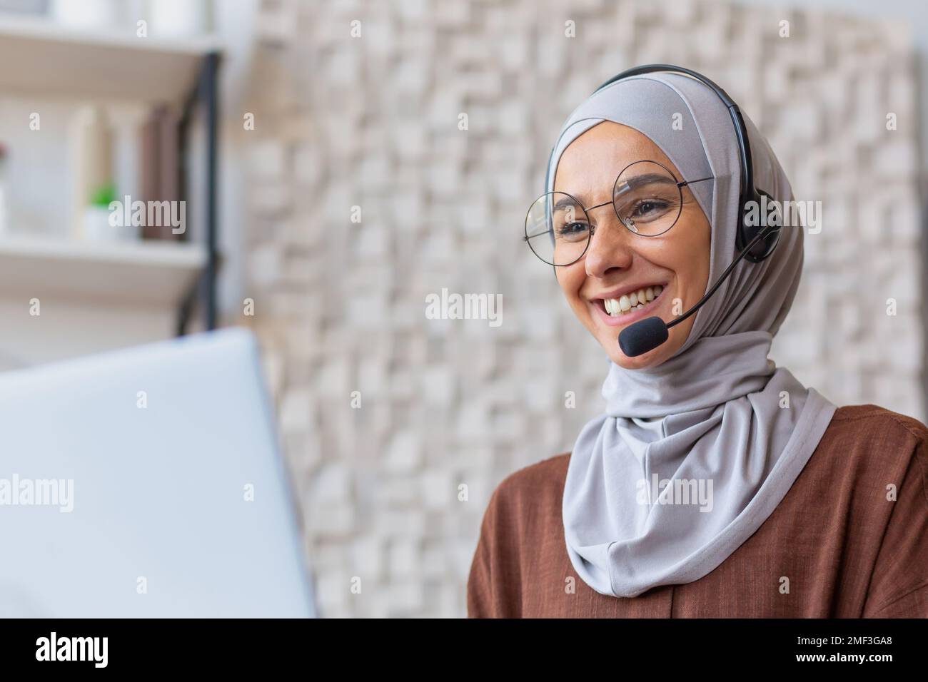 Fröhliche Frau im Hijab, die von zu Hause aus arbeitet, muslimische Frau, die mit Headset und Laptop Videoanrufe führt, Geschäftsfrau, die von zu Hause aus arbeitet. Stockfoto