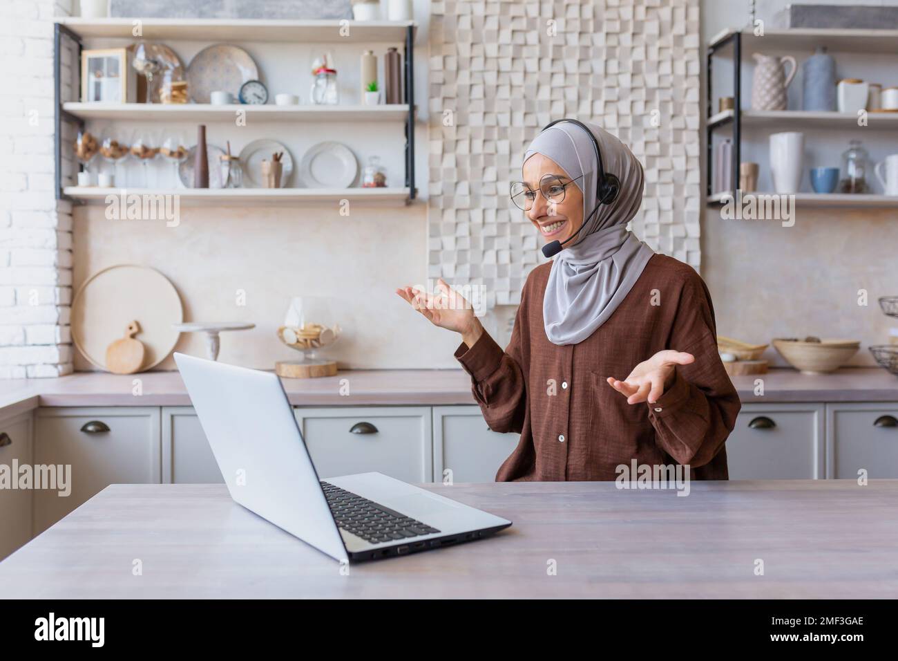 Fröhliche Frau im Hijab, die von zu Hause aus arbeitet, muslimische Frau, die mit Headset und Laptop Videoanrufe führt, Geschäftsfrau, die von zu Hause aus arbeitet. Stockfoto