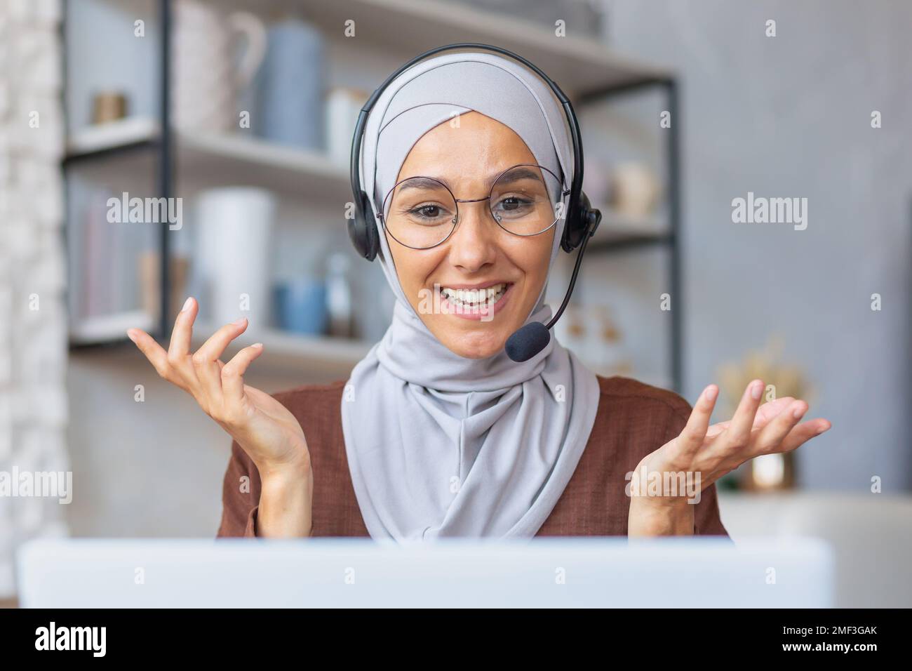 Fröhliche Frau im Hijab, die von zu Hause aus arbeitet, muslimische Frau, die mit Headset und Laptop Videoanrufe führt, Geschäftsfrau, die von zu Hause aus arbeitet. Stockfoto