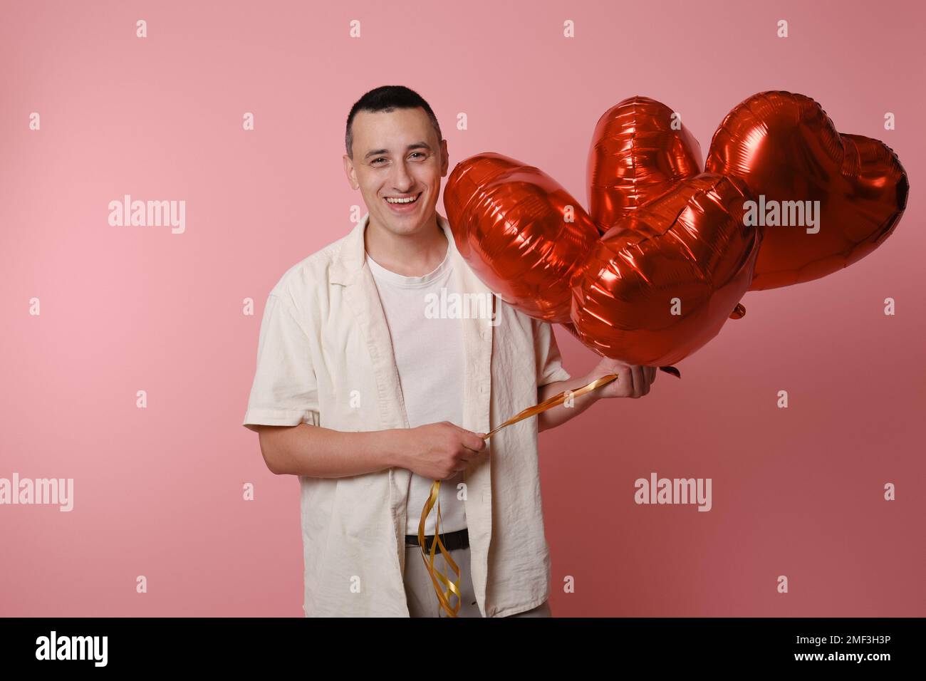 Ein Mann hält rote Ballons in Form eines Herzens auf pinkfarbenem Hintergrund. Valentinstag, Liebe, 14. Februar. Er sieht in die Kamera und lächelt Stockfoto