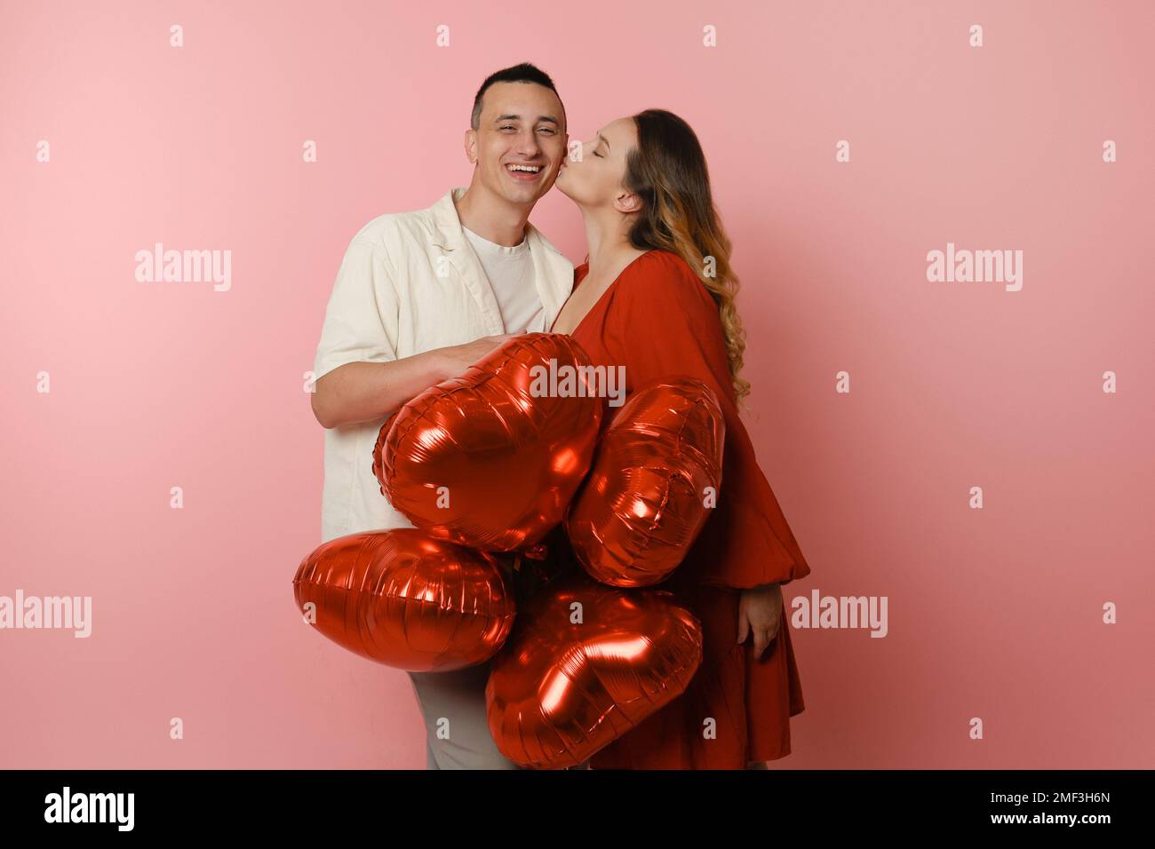 Ein Paar, rote Ballons in Form eines Herzens auf pinkfarbenem Hintergrund. Valentinstag, Liebe, 14. Februar. Eine Frau in einem roten Kleid küsst einen Mann Stockfoto