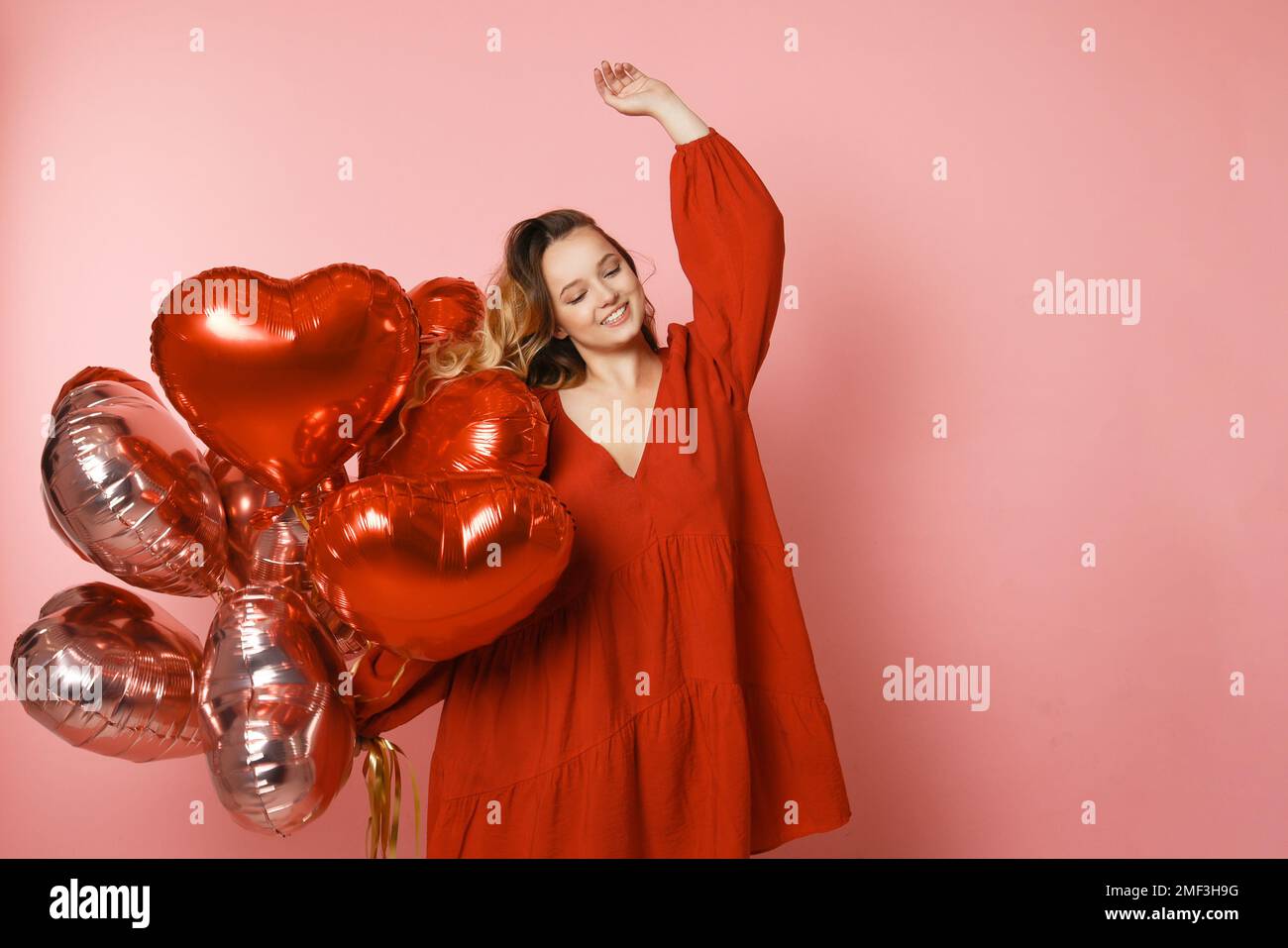 Hübsches Mädchen in einem roten Kleid auf pinkfarbenem Hintergrund, rote und pinkfarbene Herzballons. Fröhliches Model, das Spaß hat, feiert, Tänze spielt. Valentinstag Stockfoto