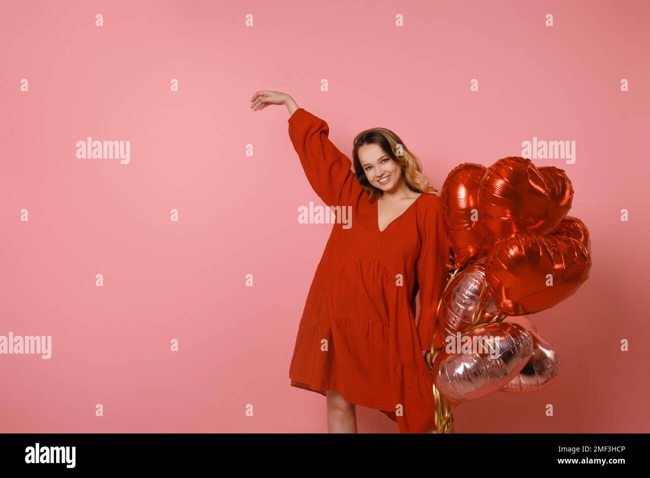 Hübsches Mädchen in einem roten Kleid auf pinkfarbenem Hintergrund, rote und pinkfarbene Herzballons. Fröhliches Model, das Spaß hat, feiert, Tänze spielt. Valentinstag Stockfoto