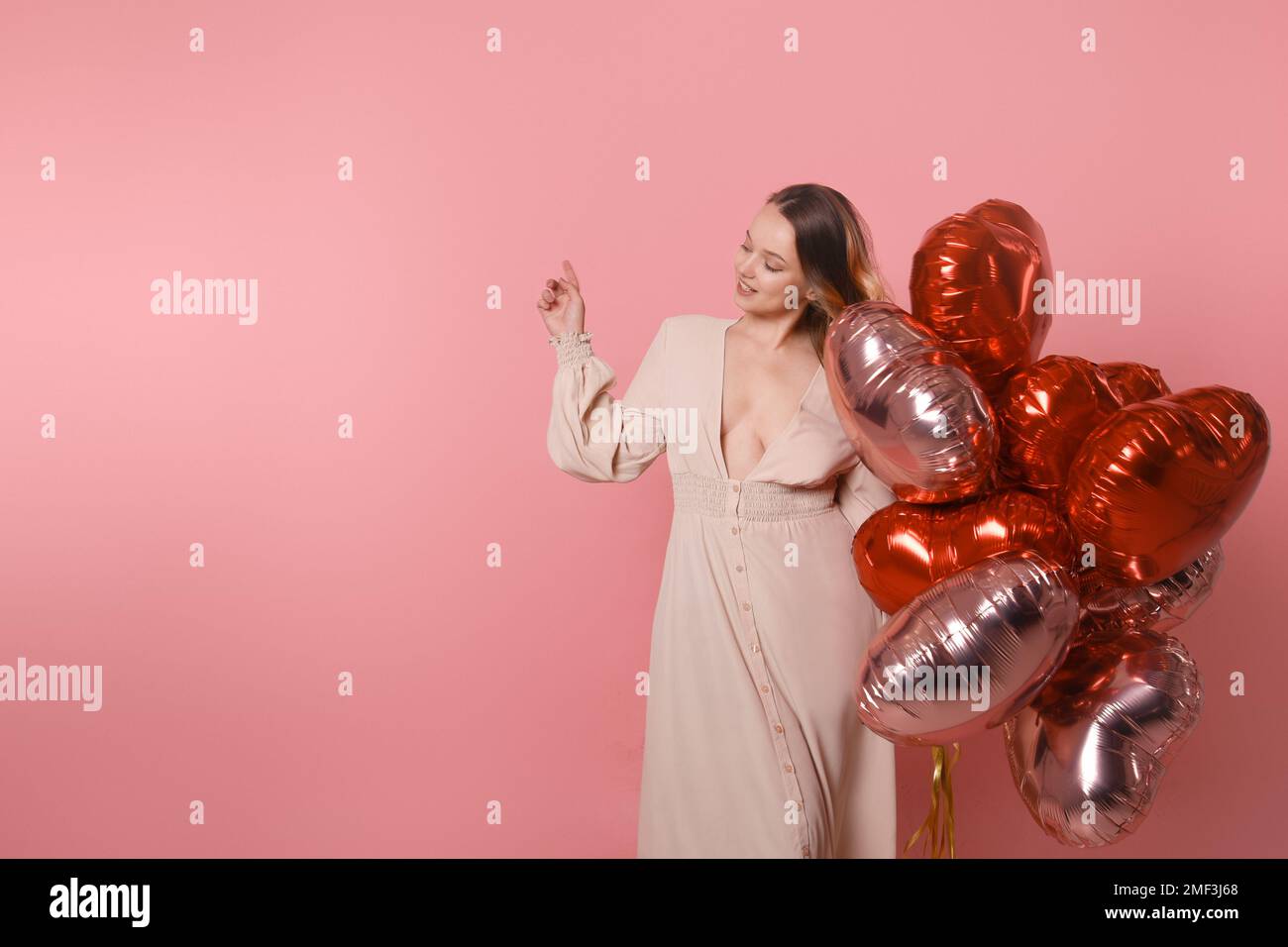 Eine Frau mit roten Ballons. Fröhliches Model, das Spaß hat, spielt und den Valentinstag feiert, in einem langen pinkfarbenen Kleid, das auf einen leeren Sitz zeigt. Überrascht, Lächeln Stockfoto