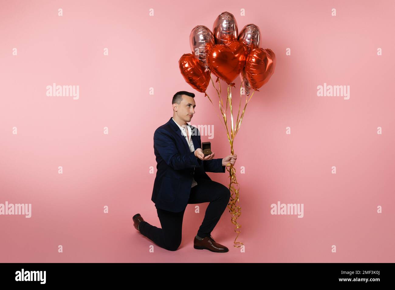 Ein Mann mit Herzballons auf pinkem Hintergrund hält eine Kiste mit Ehering. Verlobt am Valentinstag, Heiratsantrag, trauriges Gesicht, unglücklich, BA Stockfoto