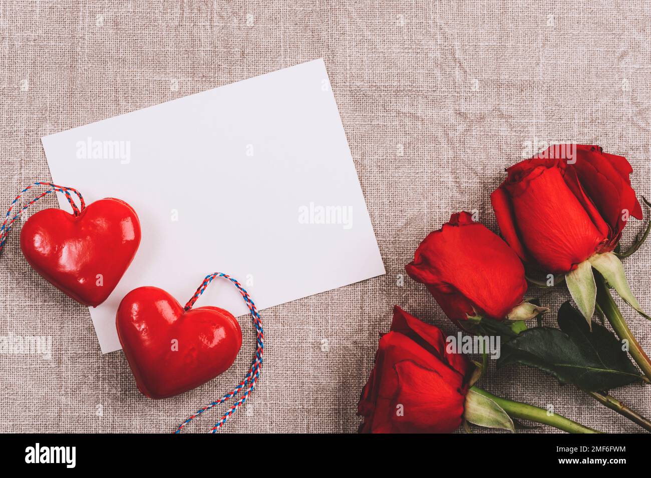 Unbeschriftete Karte, zwei Herzen und rote Rosen auf neutralem Textilhintergrund. Valentinstag-Konzept. Draufsicht, flaches Lay, Modell. Stockfoto