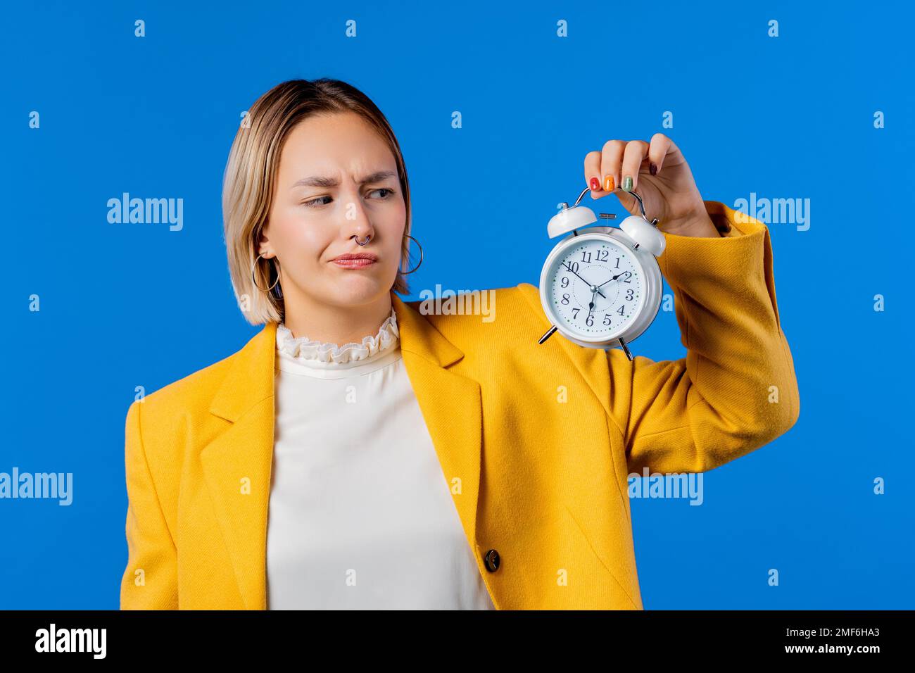 Irritierte Frau, die den Wecker in der Hand hält. Blauer Hintergrund. Früh um 6 Uhr Stockfoto