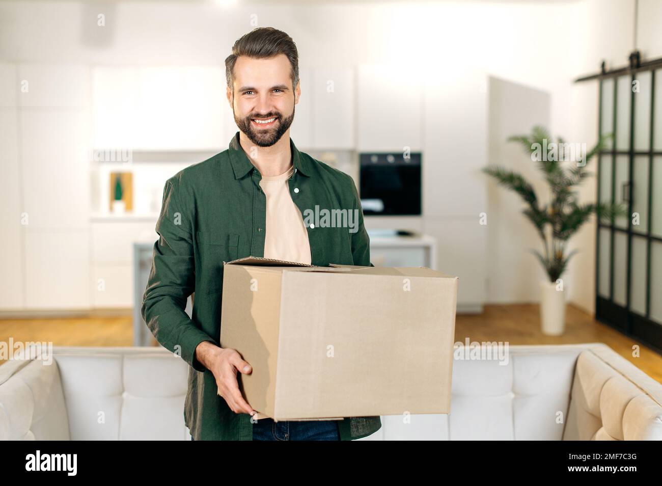 Glücklicher attraktiver, eleganter weißer Mann, stehe zu Hause im Wohnzimmer, halte einen großen Karton in den Händen, habe ein Paket aus dem Online-Shop erhalten, bereite sich auf das Auspacken vor, schaut in die Kamera, lächelt freundlich Stockfoto