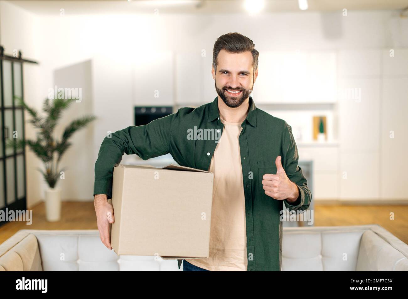 Glücklicher attraktiver, eleganter Kaukasier, zu Hause stehen, einen großen Karton in den Händen halten, ein Paket aus dem Online-Shop erhalten, sich auf das Auspacken vorbereiten, in die Kamera schauen, lächeln, Daumen-hoch-Geste Stockfoto