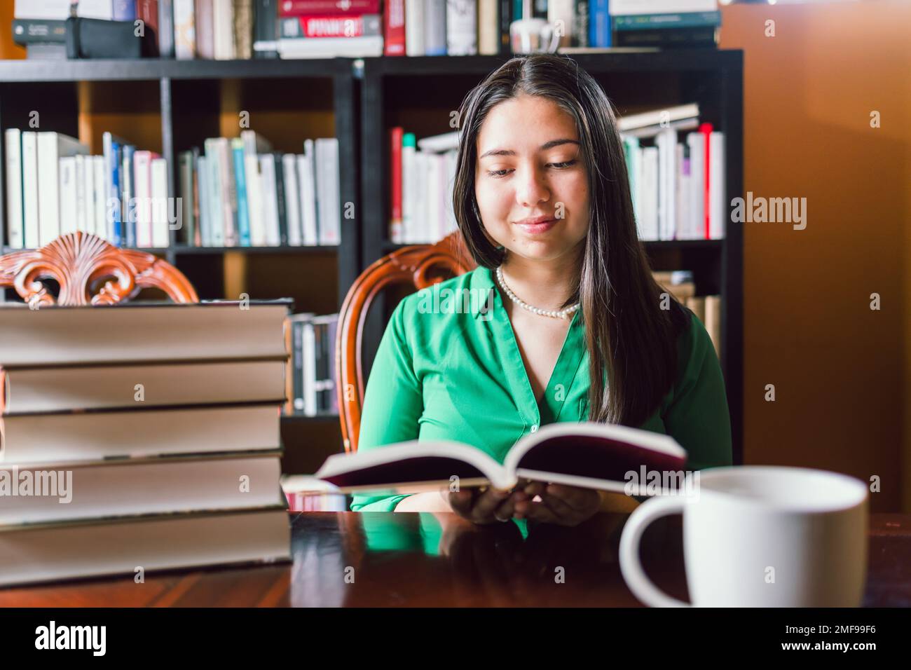 Eine junge Frau liest Bücher in ihrer persönlichen Bibliothek, mit einer Kaffeetasse. Weltbuchtag Stockfoto