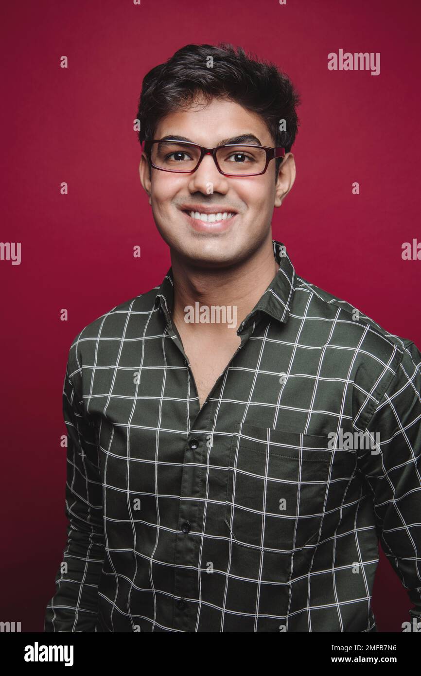 Porträt eines jungen Indianers in kariertem Hemd und Brille, der vor rotem Hintergrund vor der Kamera lächelt Stockfoto