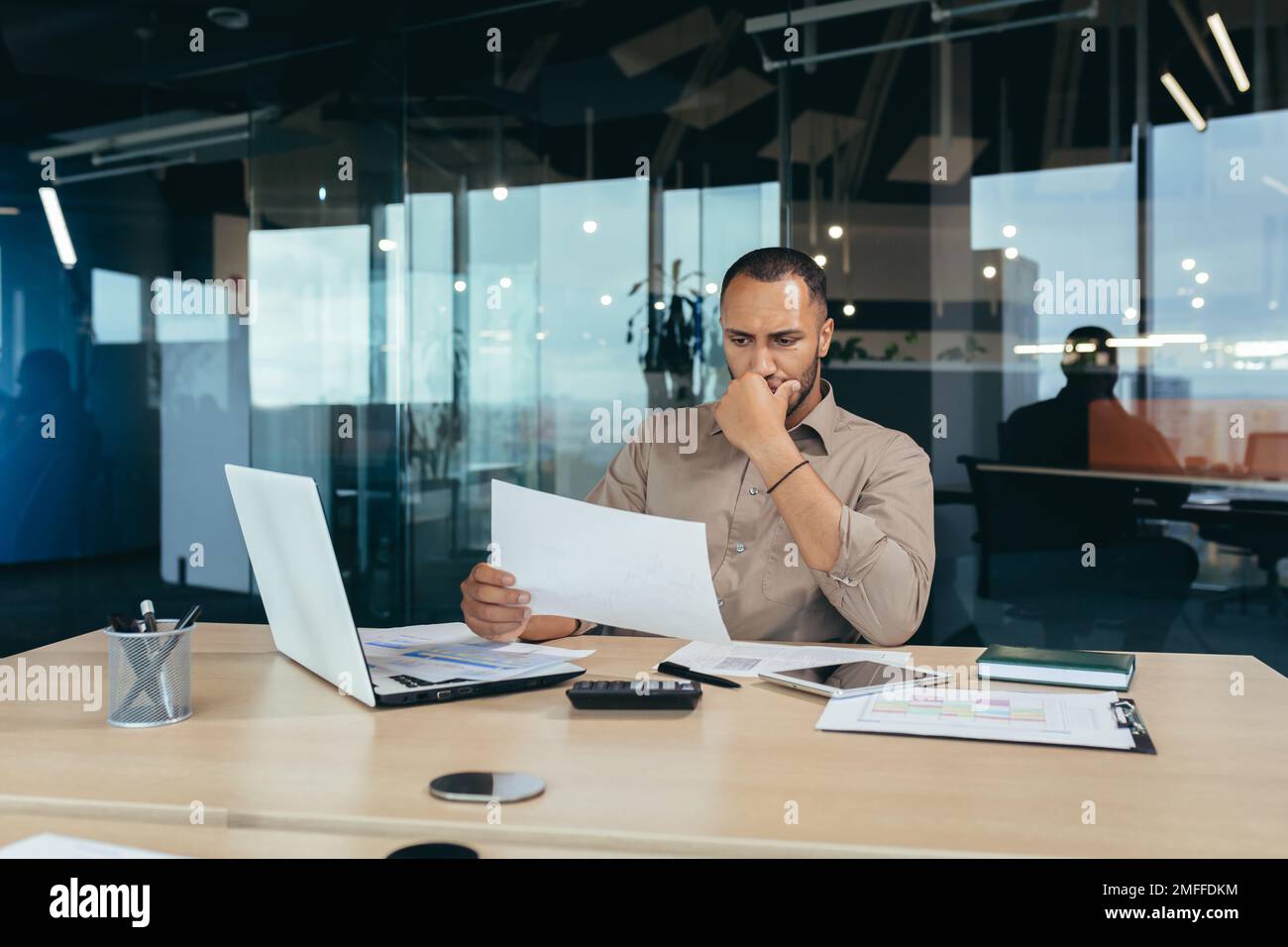 Rücksichtsvoller junger afroamerikanischer Designer, Architekt, der mit einem Laptop im Büro am Tisch sitzt. Er hält Dokumente in der Hand und konzentriert sich auf die Arbeit mit Plänen und Zeichnungen. Stockfoto