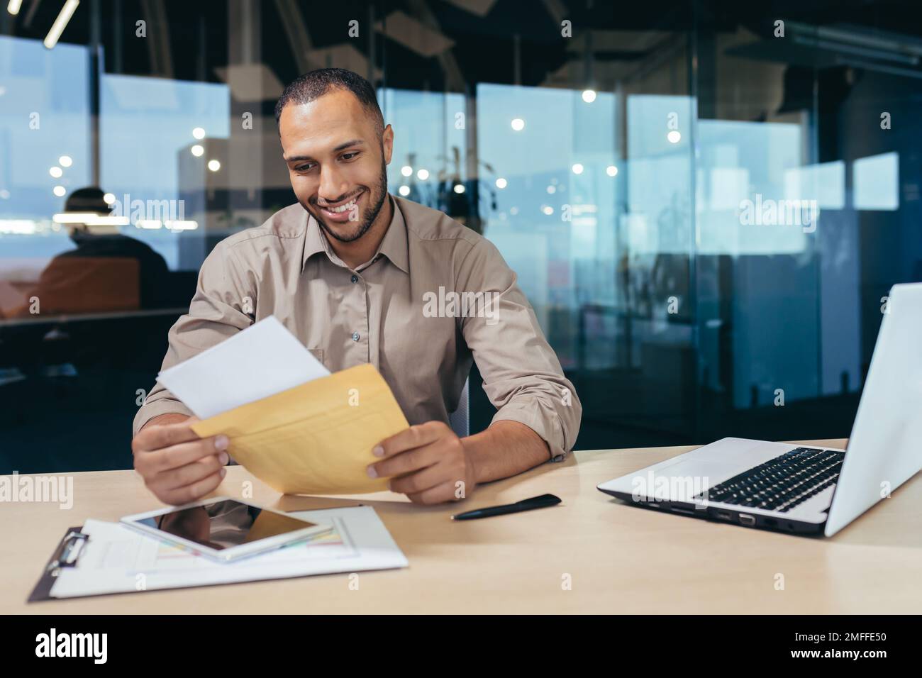 Ein glücklicher junger Mann erhielt einen Brief mit guten Nachrichten. Er sitzt im Büro an einem Tisch mit einem Laptop, hält einen Umschlag in der Hand, liest und lächelt. Stockfoto