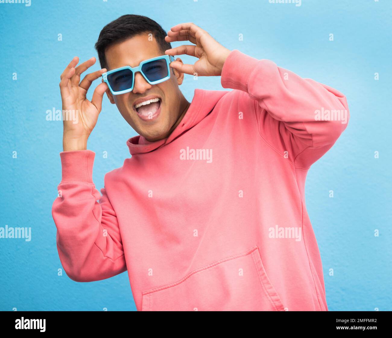 Glück, Mann mit Sonnenbrille und Lächeln mit Gesicht, trendige Mode und glücklich mit Freiheit isoliert auf blauem Hintergrund. Funky, Retro und Wellness Stockfoto
