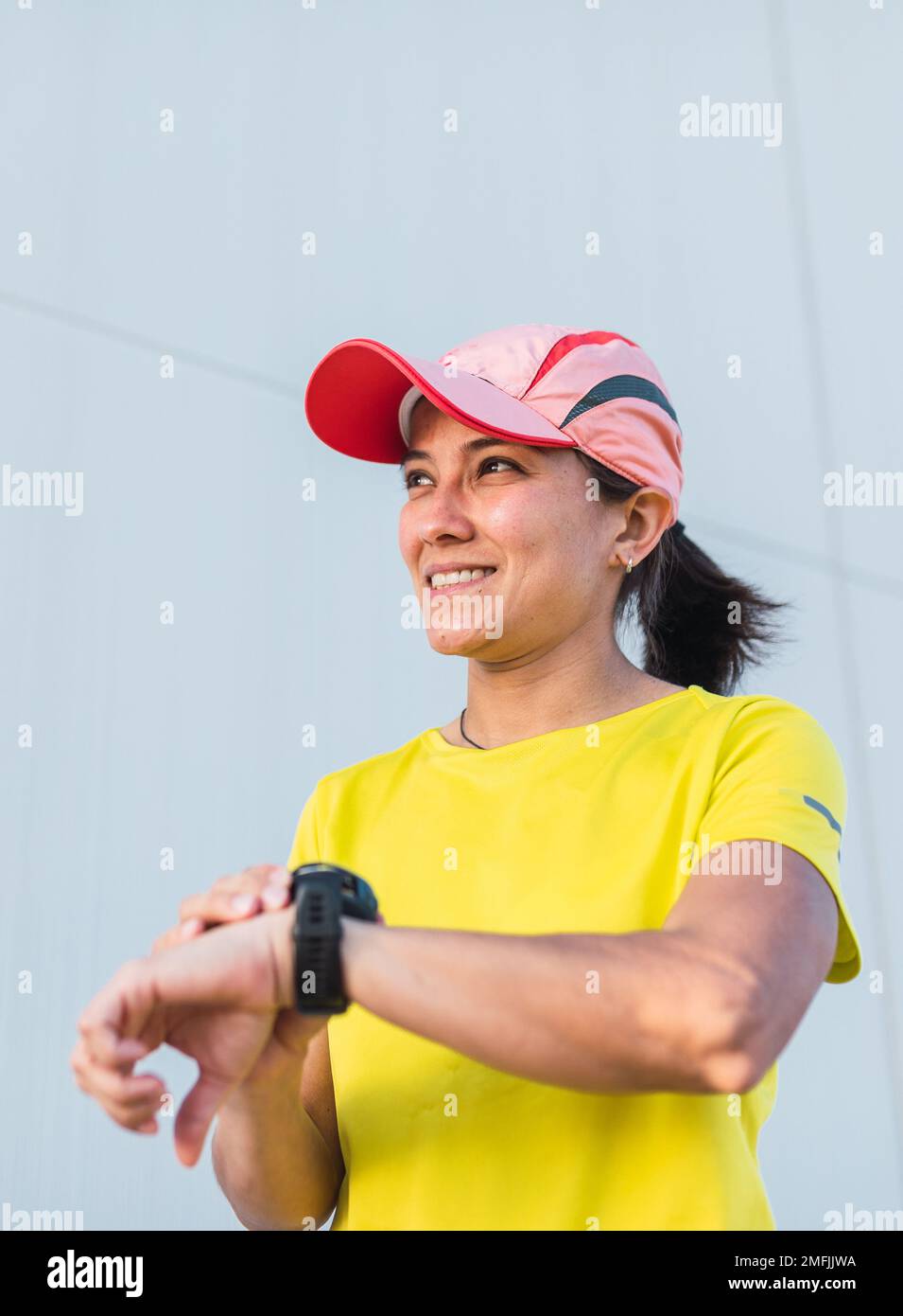 Latina-Läuferin schaut auf ihre Uhr. Sie synchronisiert ihre Uhr, um laufen zu gehen. Gelbe Kleidung und Mütze. Stockfoto