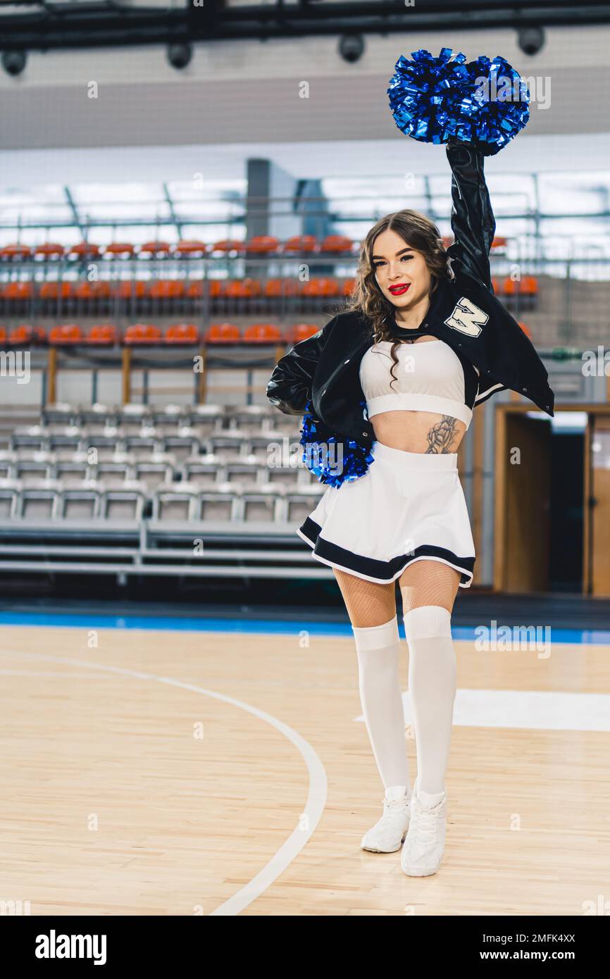 Vertikale Aufnahme einer Cheerleaderin in Uniform mit schwarzer Jacke und weißem Rock, der auf dem Basketballfeld posiert. Die Boxen der Sporthalle verschwommen im Hintergrund. Hochwertiges Foto Stockfoto