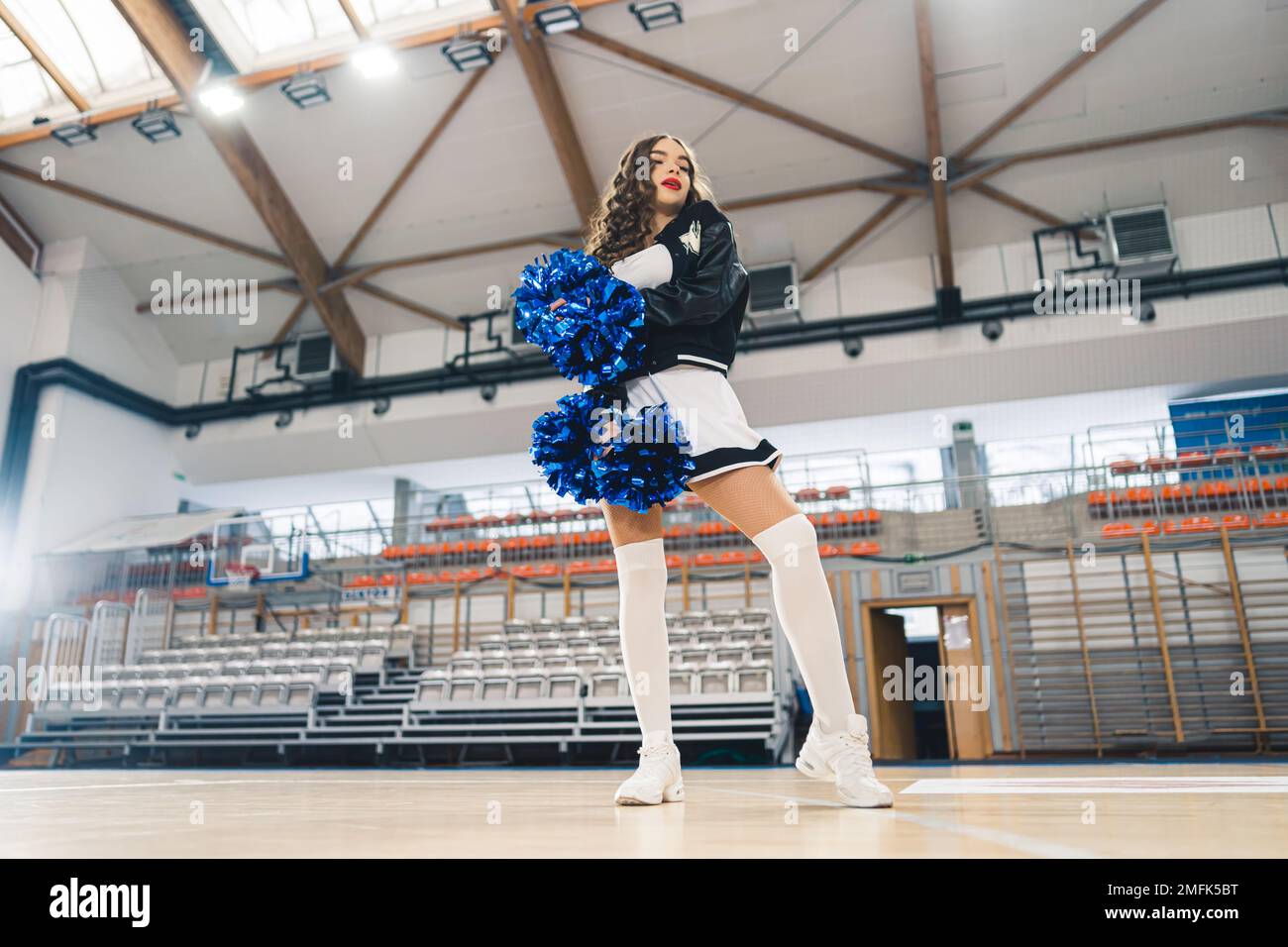 Horizontale Aufnahme der braunen Cheerleaderin in Schwarz-Weiß-Uniform mit blauen, glänzenden Bommeln von unten. Basketballfeld im Hintergrund. Hochwertiges Foto Stockfoto