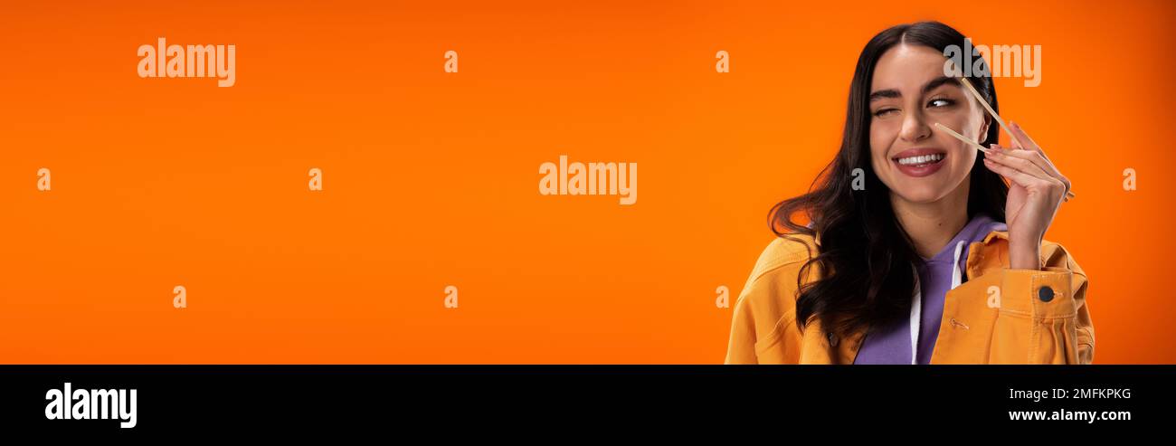 Junge Frau, die Bambusstäbchen in der Hand hält und isoliert zwinkert, auf Orange, Banner, Stock Image Stockfoto
