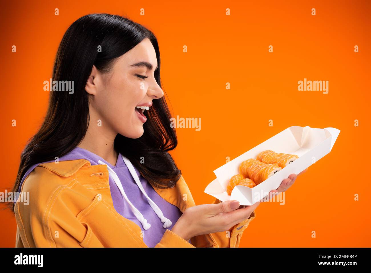 Seitenansicht einer fröhlichen Frau, die sich Sushi zum Mitnehmen ansah, isoliert auf einem orangefarbenen Stockbild Stockfoto