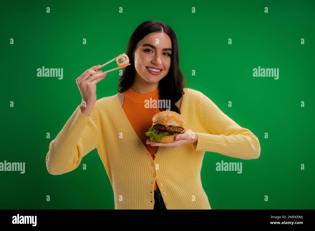Glückliche junge Frau mit Burger und Essstäbchen mit Sushi-Rolle isoliert auf grünem Stock Image Stockfoto