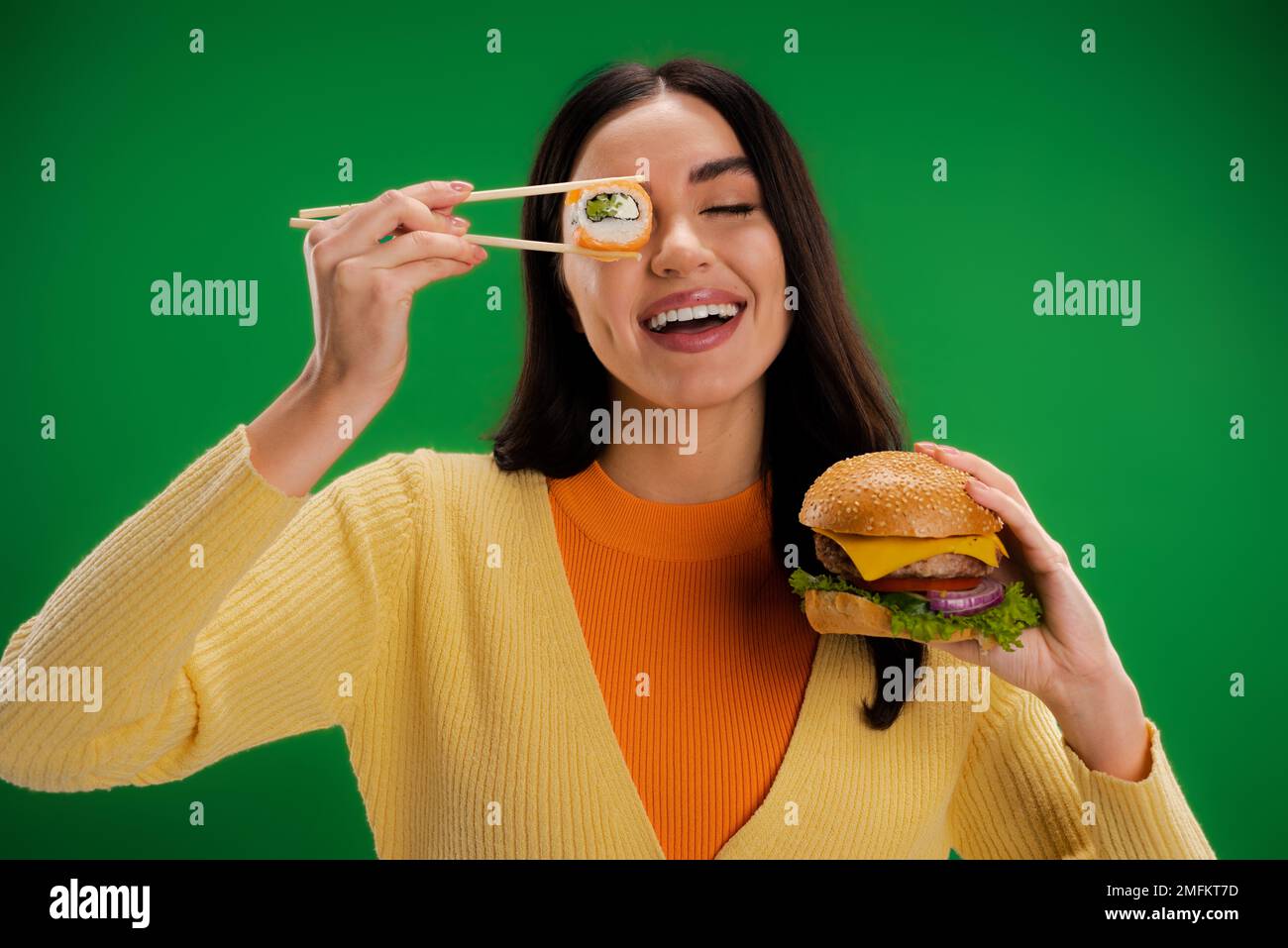 Fröhliche Frau mit leckeren Burgern, die Sushi-Rollen nahe dem Auge hält, isoliert auf grünem Stockbild Stockfoto