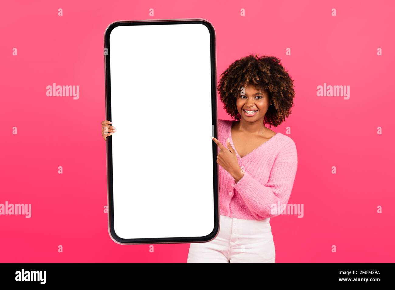 Fröhlich hübsche junge afroamerikanische lockige Frau in lässiger Hosenträger halten großes Smartphone mit leerem Bildschirm Stockfoto