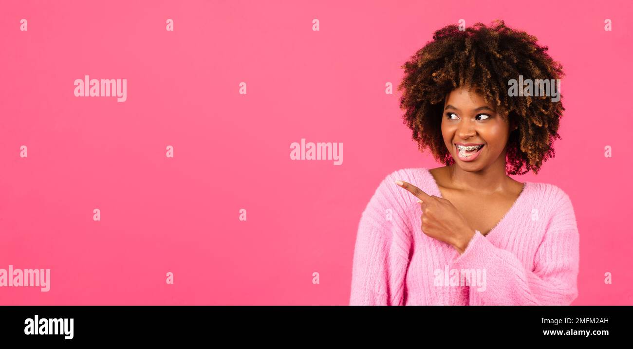 Eine glückliche, afroamerikanische, lockige Millenial-Frau in ungezwungener Form, mit offenem Mund, Finger auf freien Platz Stockfoto