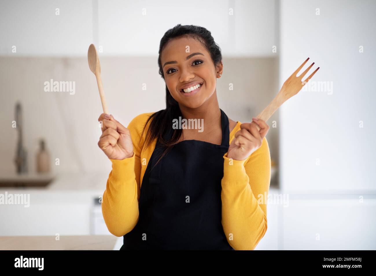 Eine fröhliche, afroamerikanische Chefköchin mit Holzlöffel und Gabel in der Schürze, die Essen kocht Stockfoto