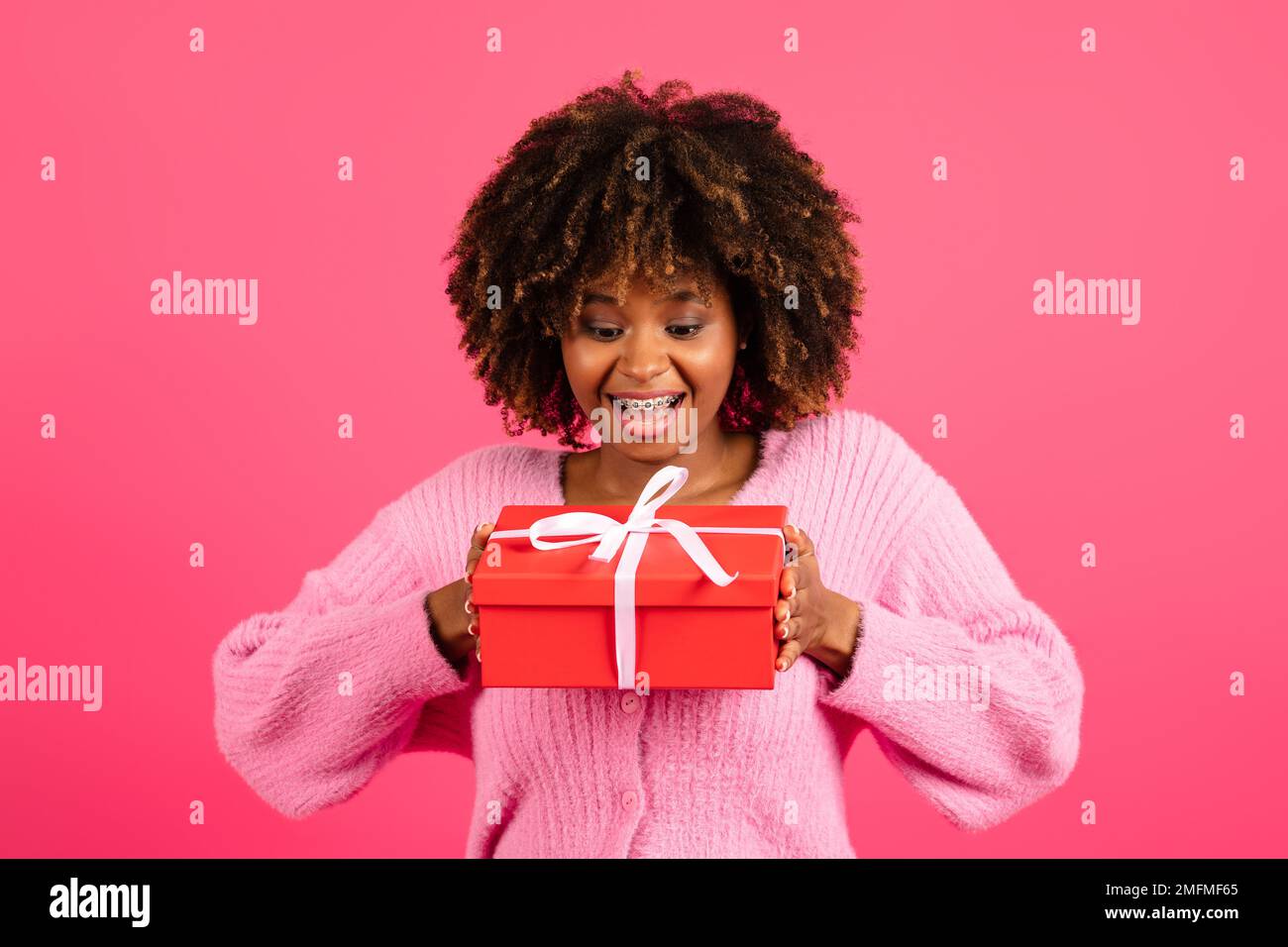 Fröhliche, schockierte junge, schwarze, lockige Dame in lässiger Zahnspange sieht auf die Schachtel mit Geschenk Stockfoto