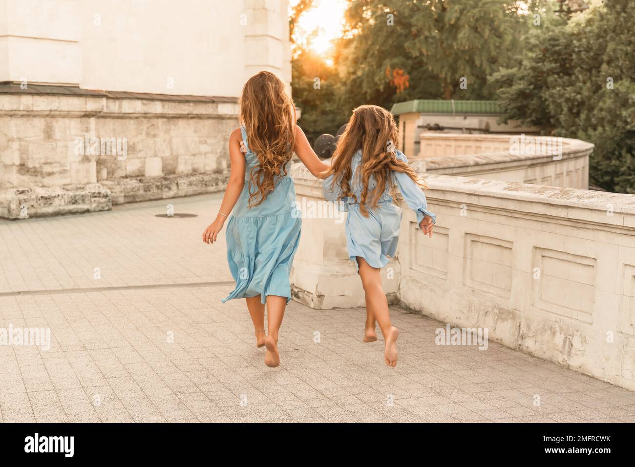 Tochter Mutter lief und hielt Händchen. In blauen Kleidern mit fließenden langen Haaren vor dem Hintergrund eines Sonnenuntergangs und einem weißen Gebäude. Stockfoto