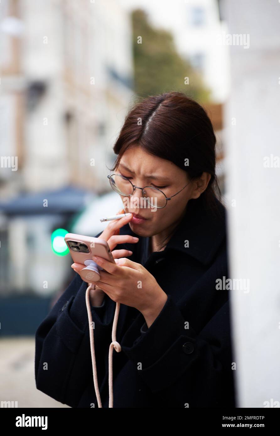 Frau raucht, während sie auf den Straßen des Stadtzentrums auf ihr Smartphone schaut Stockfoto