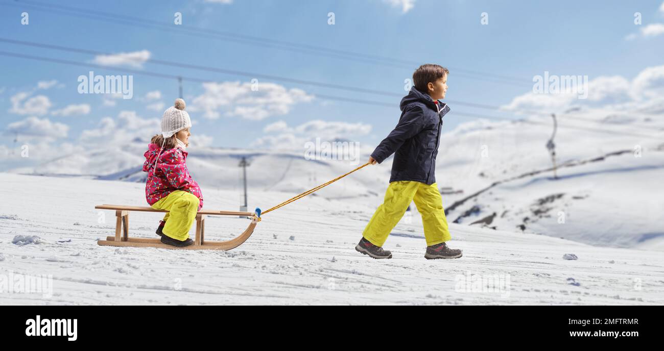 Profilaufnahme über die gesamte Länge eines Jungen, der ein Mädchen auf einem hölzernen Schlitten auf einem verschneiten Berg zieht Stockfoto