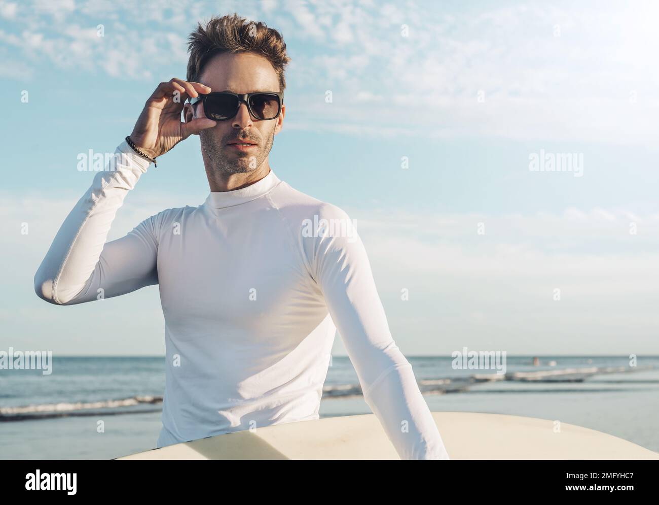 Junger, gutaussehender Surfer, der ein leeres Lycra-T-Shirt trägt und ein Surfbrett in der Hand hat. Sonnenlicht. Surfstil. Stockfoto