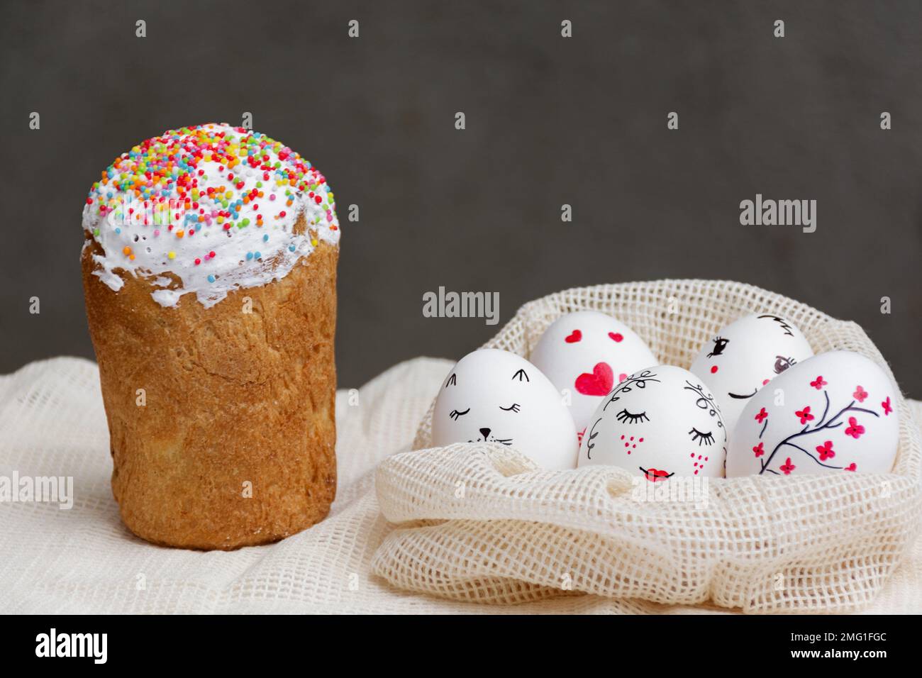 Köstlicher kulichkuchen, dekoriert mit süßem Zucker. Weiße Ostereier in einem Ökobeutel aus Netzgewebe mit wunderschönen bemalten Gesichtern auf dunklem Hintergrund. Frohe Ostern. Stockfoto