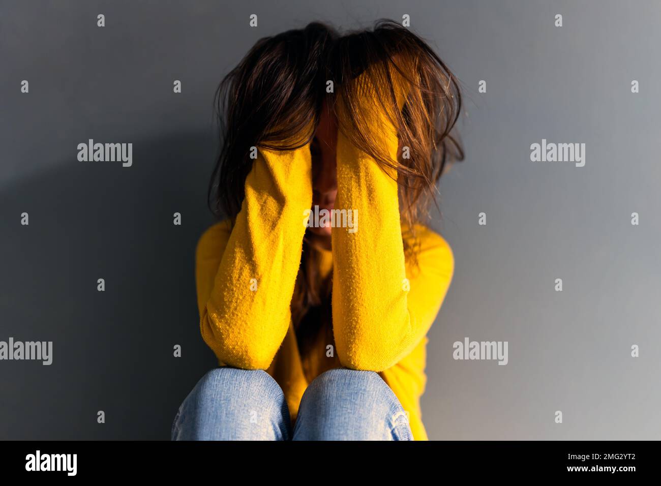 Verärgerte junge deprimierende Frau in Jeans und Pullover, die langes braunes Haar berührte, während sie auf dem Boden saß und sich an eine graue Wand lehnte Stockfoto