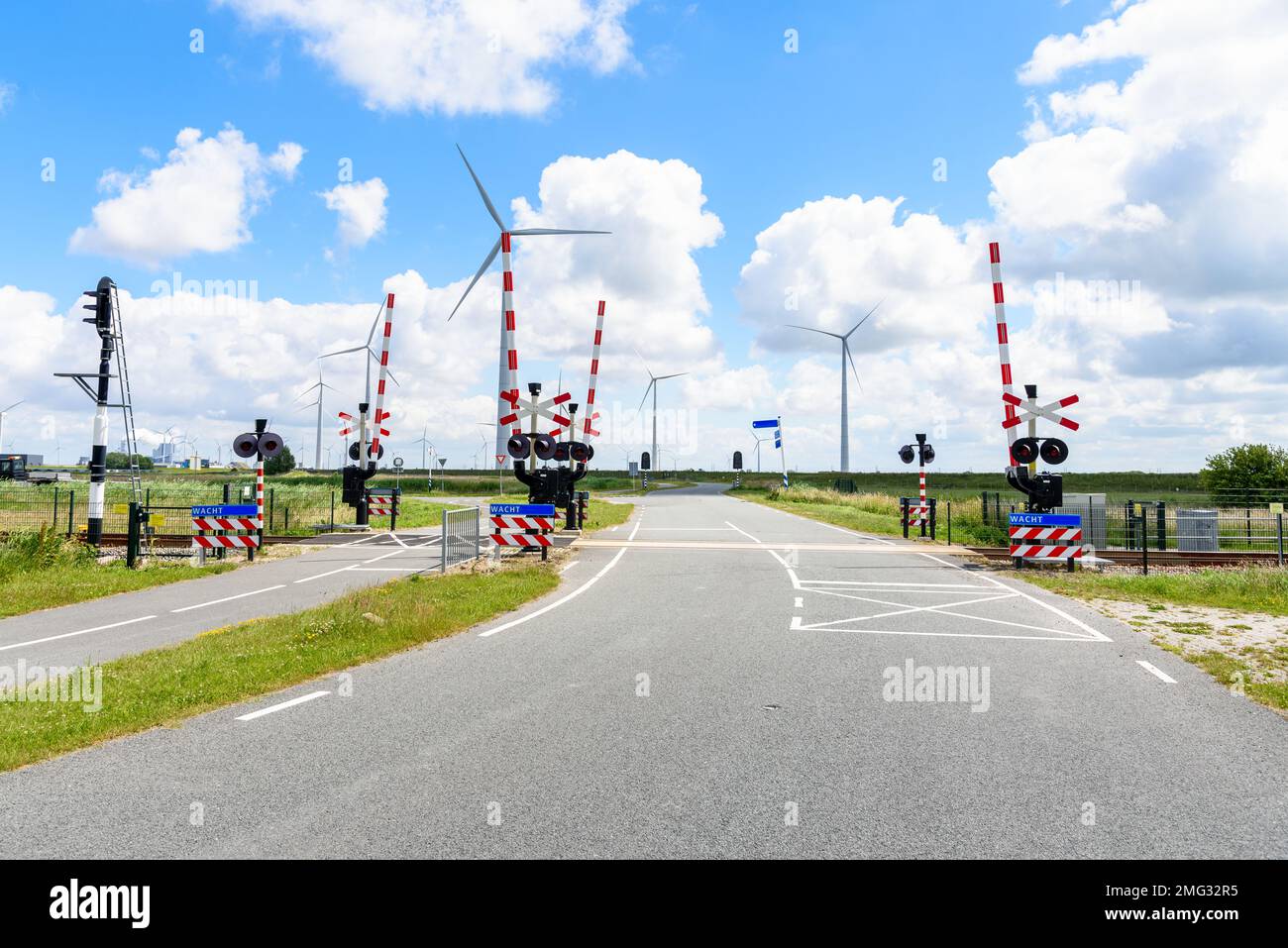 Bahnübergang mit Signalen und Warnschildern entlang einer Straße und einer Fahrradspur auf dem Land der Niederlande. Ein Windpark befindet sich im Hintergrund. Stockfoto