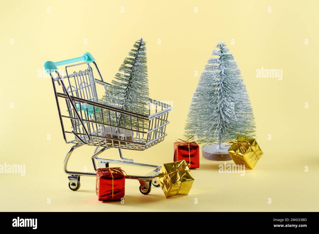 Kleiner Weihnachtsbaum in einem kleinen Einkaufswagen. Ein weiterer Weihnachtsbaum mit bunten Geschenken ist im Hintergrund. Weihnachtseinkaufskonzept. Speicherplatz kopieren Stockfoto