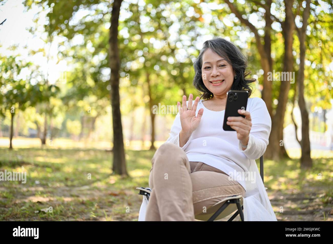 Glückliche und fröhliche Asiatin im Alter von 60s Jahren in bequemen Kleidern, die sich mit ihrem Enkelkind per Videoanruf unterhält, mit der Hand winkt, um Hallo zu sagen, sich im Park entspannt. Stockfoto