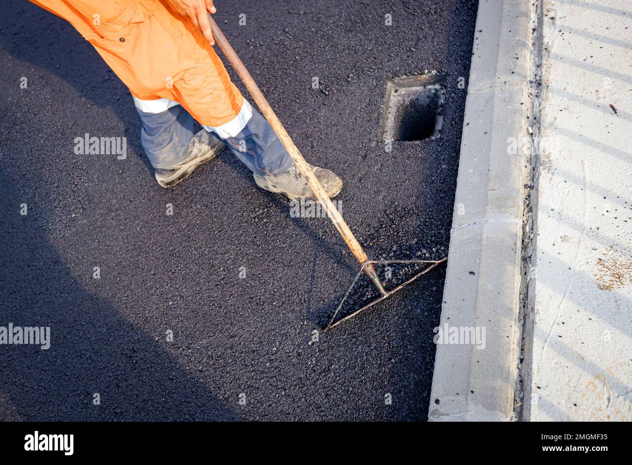 Über Sicht auf einen Arbeiter, der Rechen verwendet, um zu nivellieren, eine Schicht frischen Asphalt nach den richtigen Maßstäben aufzustellen, flacht den heißen Asphalt um das quadratische Regenwasser ab Stockfoto