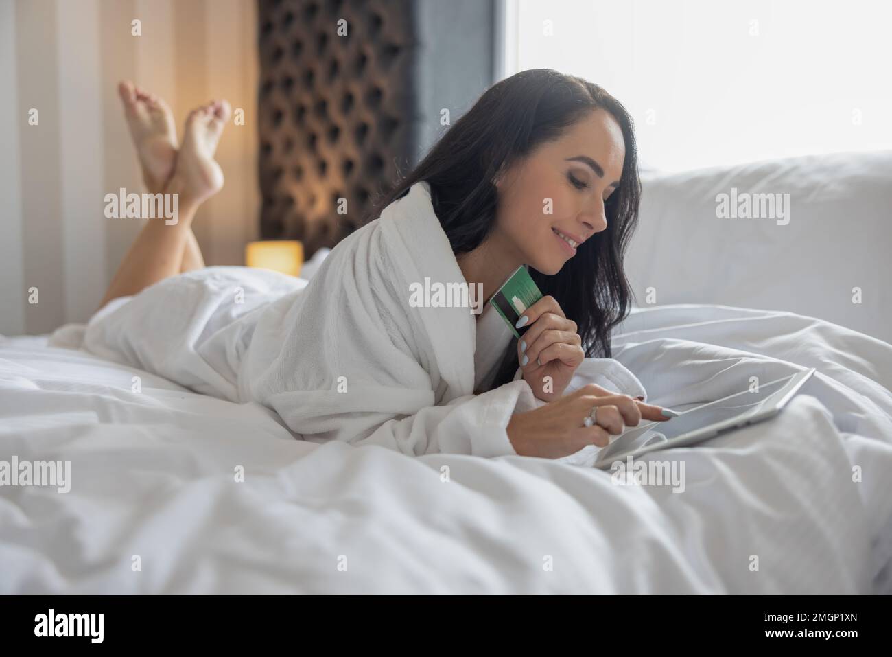 Eine lächelnde, gut aussehende Brünette aus dem Komfort ihres Schlafzimmers bestellt Dinge online auf einem Tablet und hält eine Kreditkarte. Stockfoto