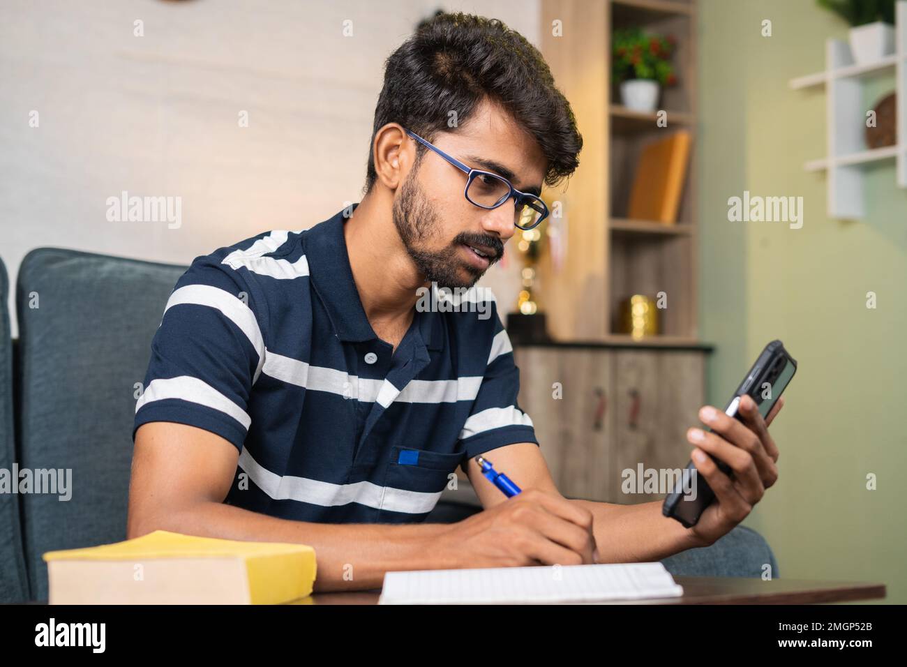 Junger Mann, der zu Hause mit dem Handy Notizen schreibt, während er Online-Kurs hört - Konzept von E-Learning, Hingabe und Intelligenz. Stockfoto