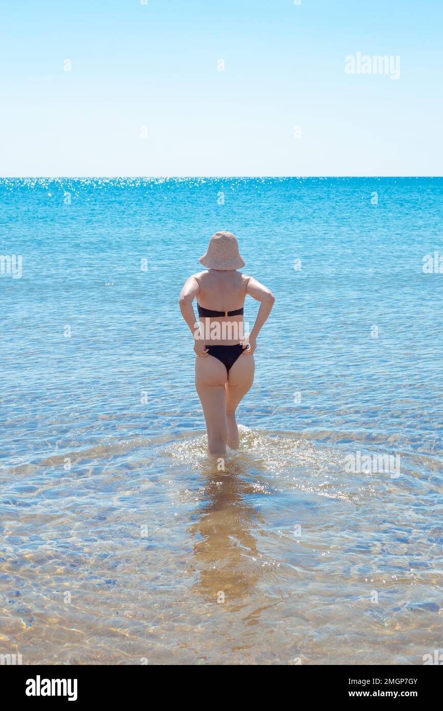 Eine reife Frau mit einer schlanken Figur im Hut geht ins Meer. Sommerurlaub an der Küste. Reisen und Tourismus. Stockfoto