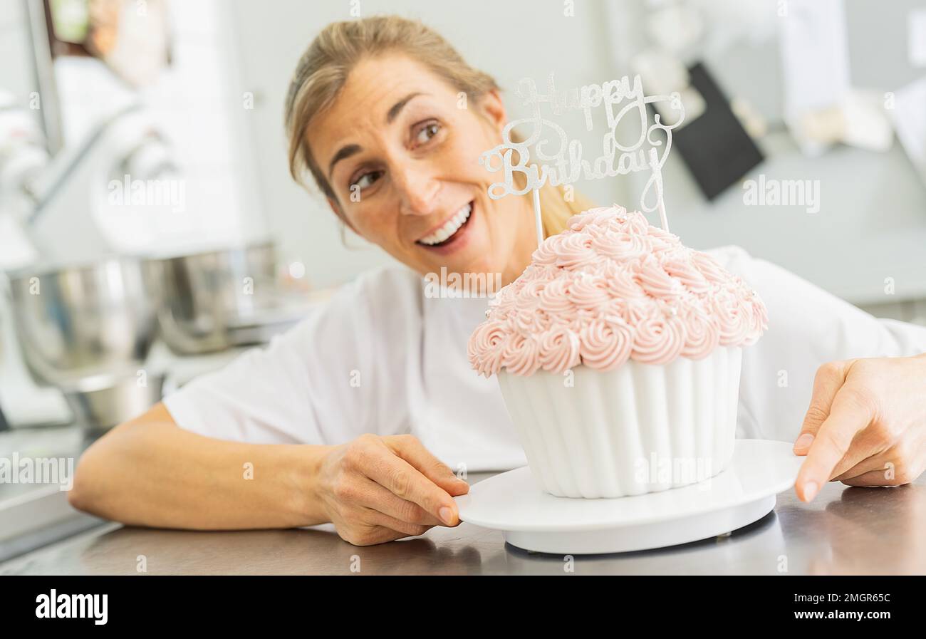 Fröhliche Konditorin lächelt und hält einen großen Geburtstagskuchen-Muffin mit Süßigkeiten in der Bäckerei Stockfoto