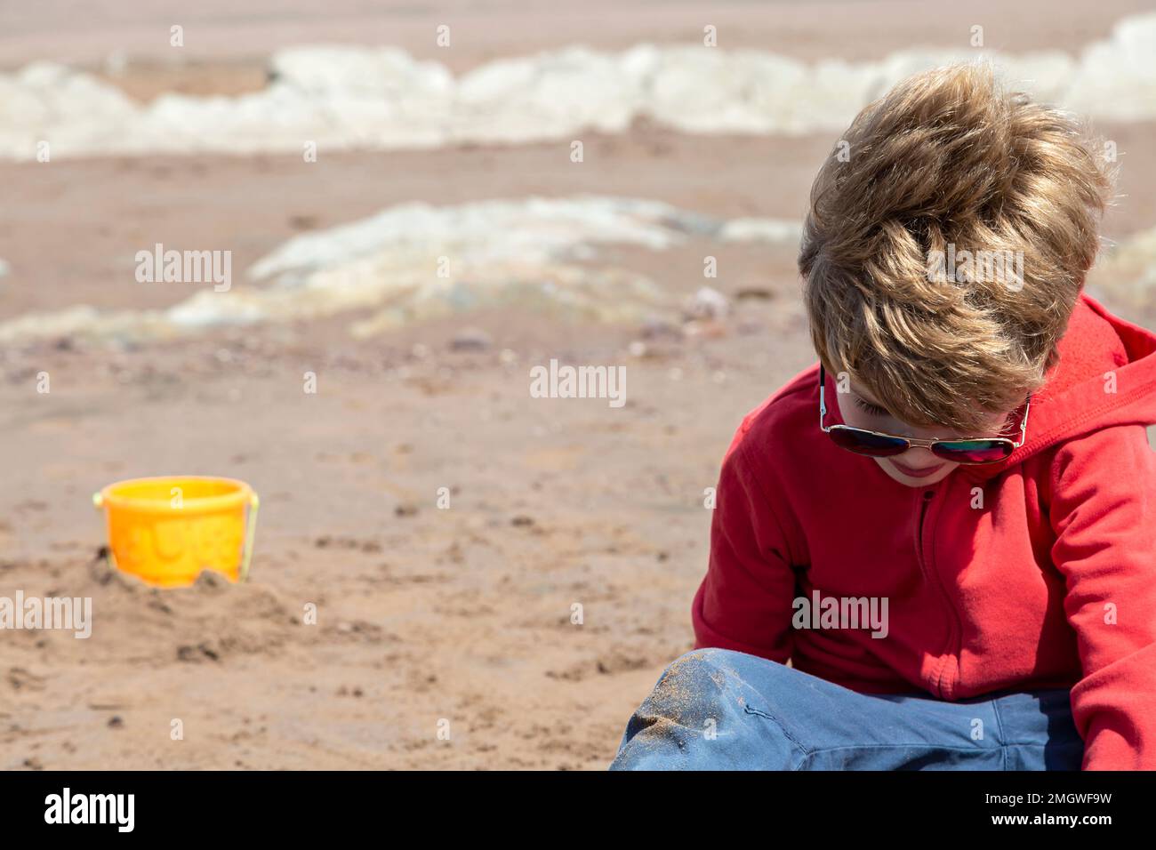Ein 7-jähriger Junge sitzt am Sandstrand von Blue Anchor, Somerset, Großbritannien Stockfoto