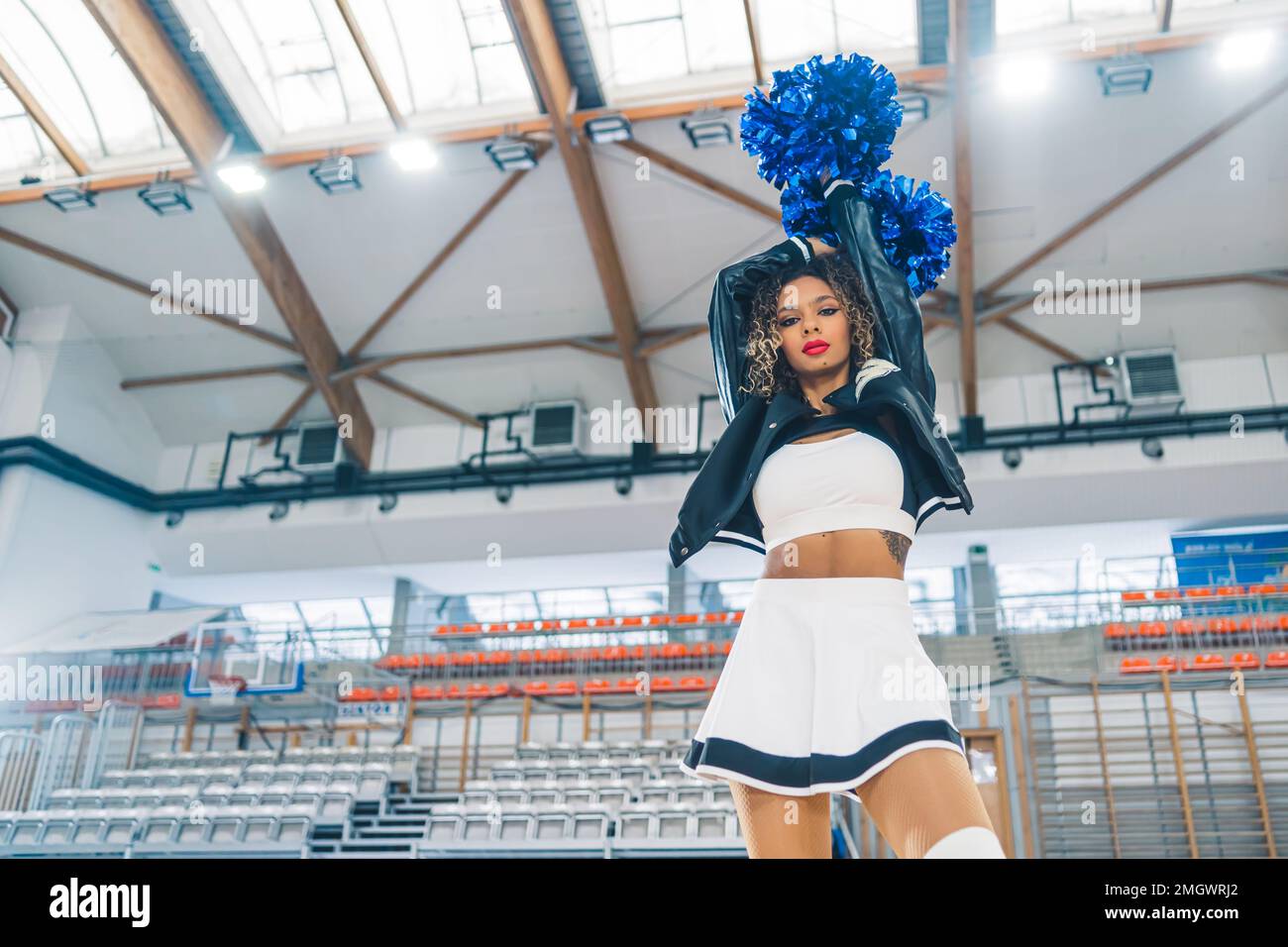 Ansicht von unten auf eine Cheerleaderin mit Lederjacke, die blaue Bommel über ihrem Kopf hält. Verschwommenes Basketballfeld im Hintergrund. Hochwertiges Foto Stockfoto