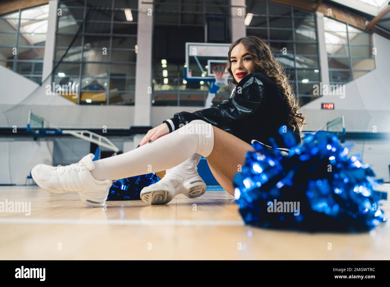 Horizontale Vollaufnahme einer Cheerleaderin schwarz-weißer Uniform, die auf dem Basketballplatz sitzt. Blau glänzender Pom-Pom verschwommen im Vordergrund. Hochwertiges Foto Stockfoto