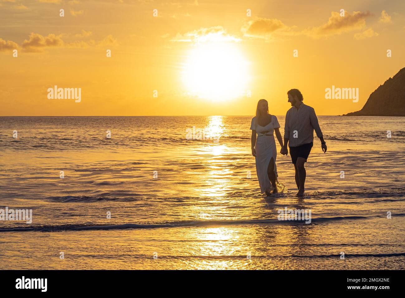 Verkleidetes junges romantisches Paar, das auf einem tropischen Sandstrand während des Sonnenuntergangs über dem Meer spaziert Stockfoto