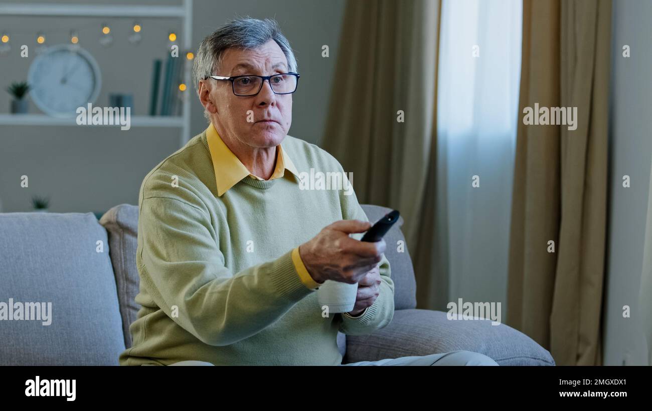 Weißer alter alter alter alter Mann in Brille entspannen Sie sich auf der Couch zu Hause, wechseln Sie den Sender mit Fernbedienung und sehen Sie sich ein Fernsehprogramm an, entspannen Sie sich beim Trinken Stockfoto