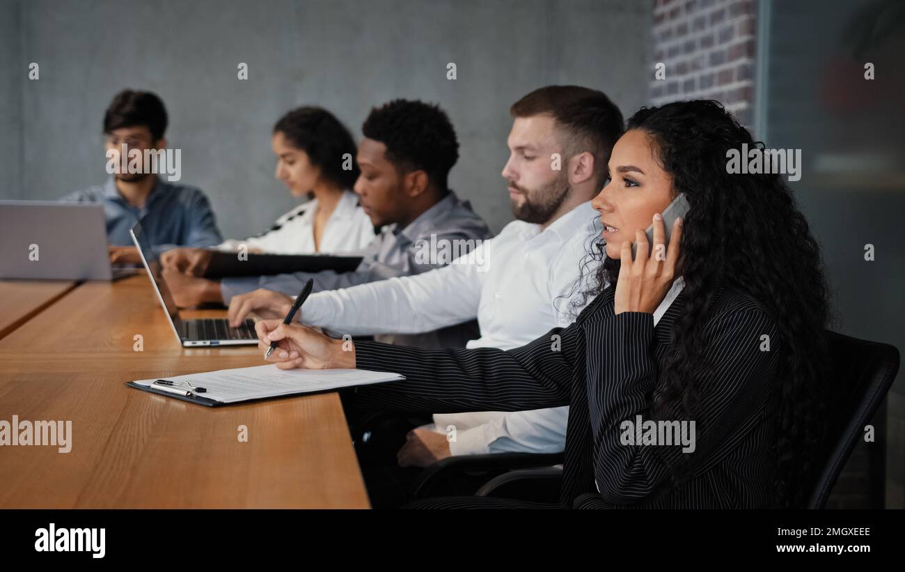 Mitarbeiter verschiedener Rassen, die im Konferenzraum sitzen, arbeiten am Tisch zusammen. Hispanische Geschäftsfrau, CEO-Managerin, die am Handy spricht und Notizen schreibt Stockfoto