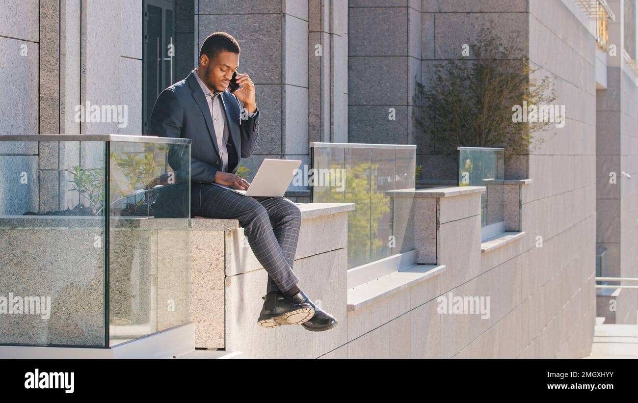Afroamerikanischer Geschäftsmann Angestellter ethnischer Unternehmer Freiberufler Verkäufer Arbeitgeber, der mit einem Laptop im Freien arbeitet und über Smartphone-Geschäft spricht Stockfoto